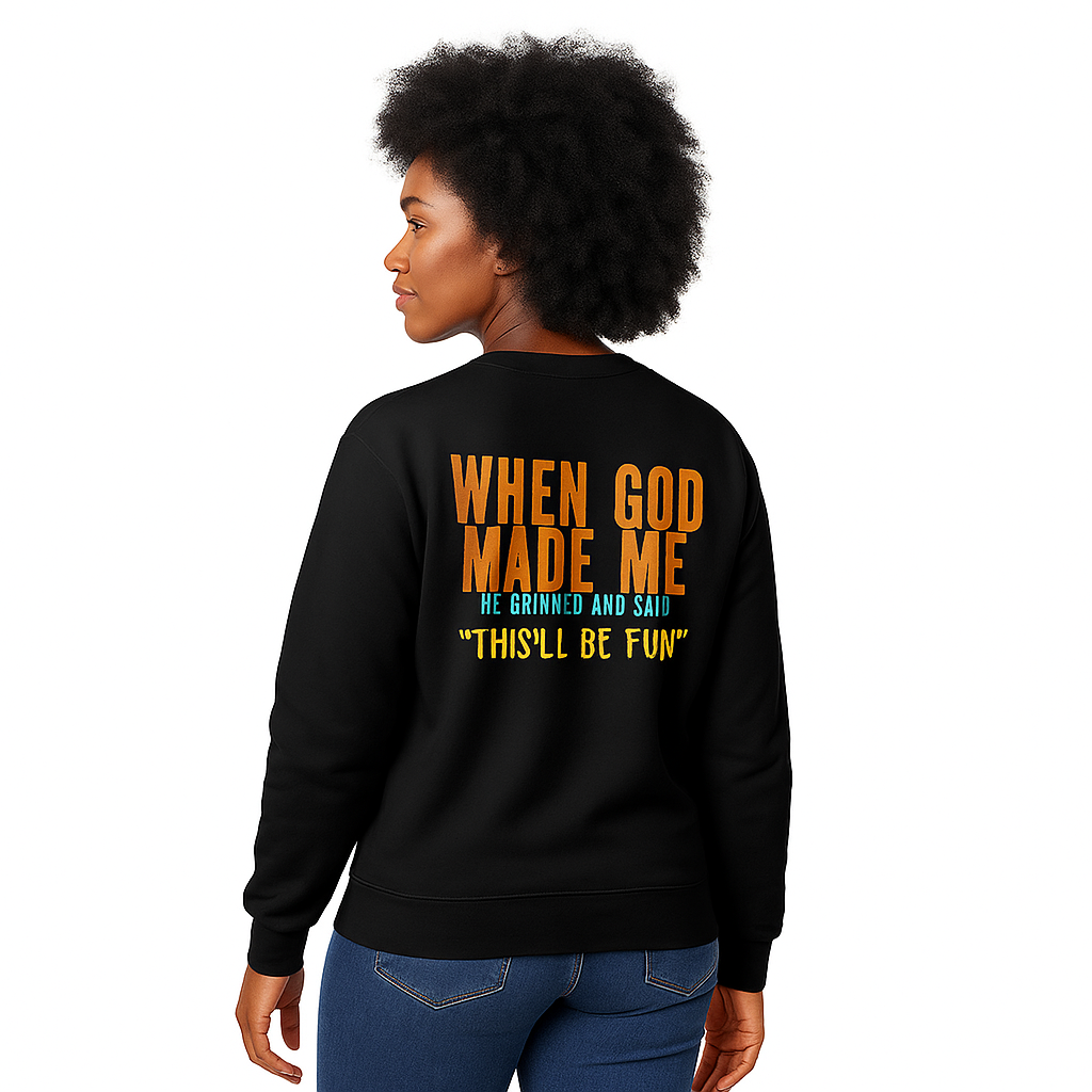 Female model wearing a black unisex crewneck sweatshirt with orange faith and humor quote When God Made Me He Grinned and Said This’ll Be Fun