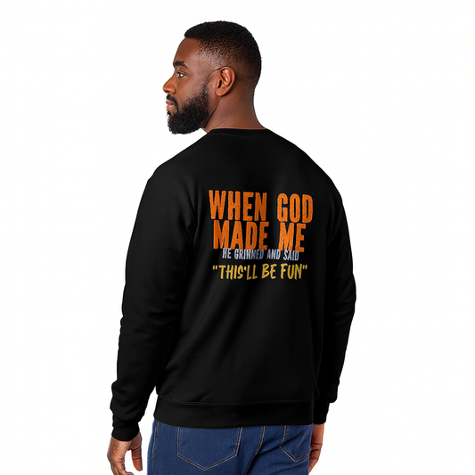 Male model wearing a black unisex crewneck sweatshirt with orange faith and humor text reading When God Made Me He Grinned and Said This’ll Be Fun